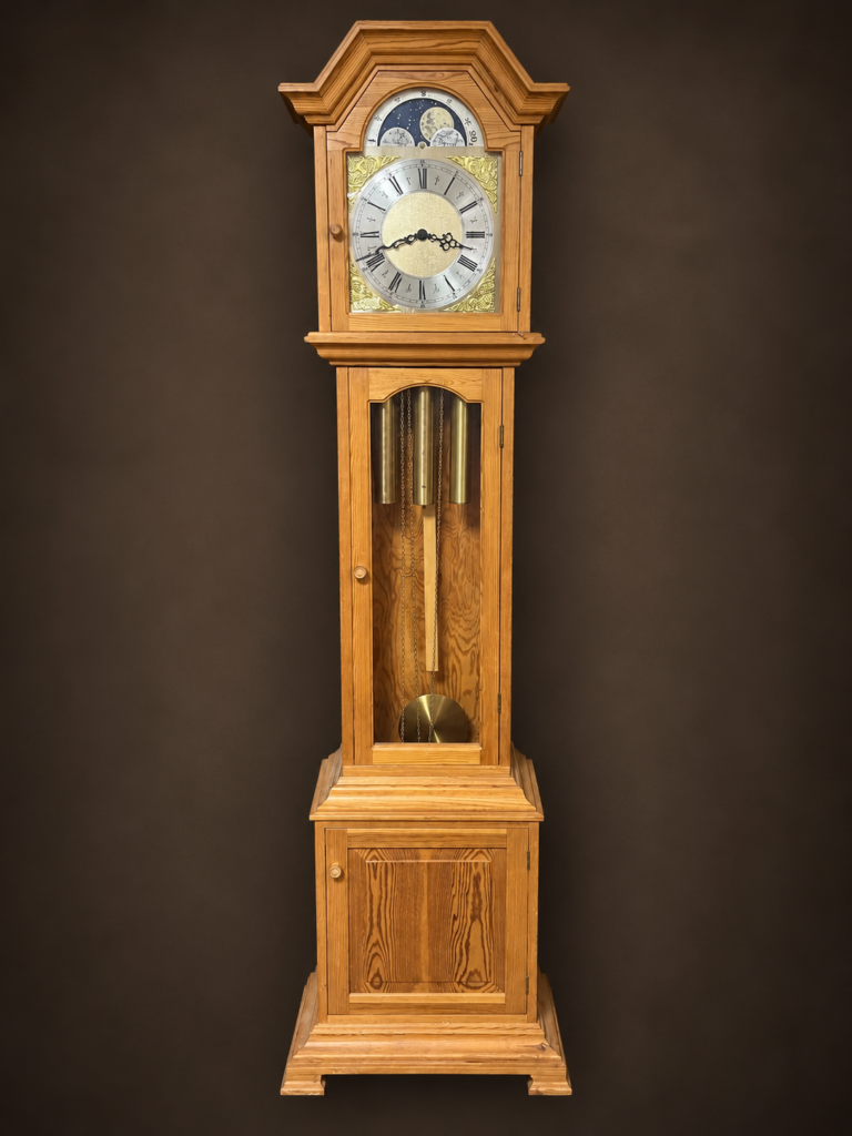 Vintage light oak grandfather clock with ornate Roman numeral dial, moon phase display, three brass weights, glass pendulum compartment, and lower wooden cabinet.