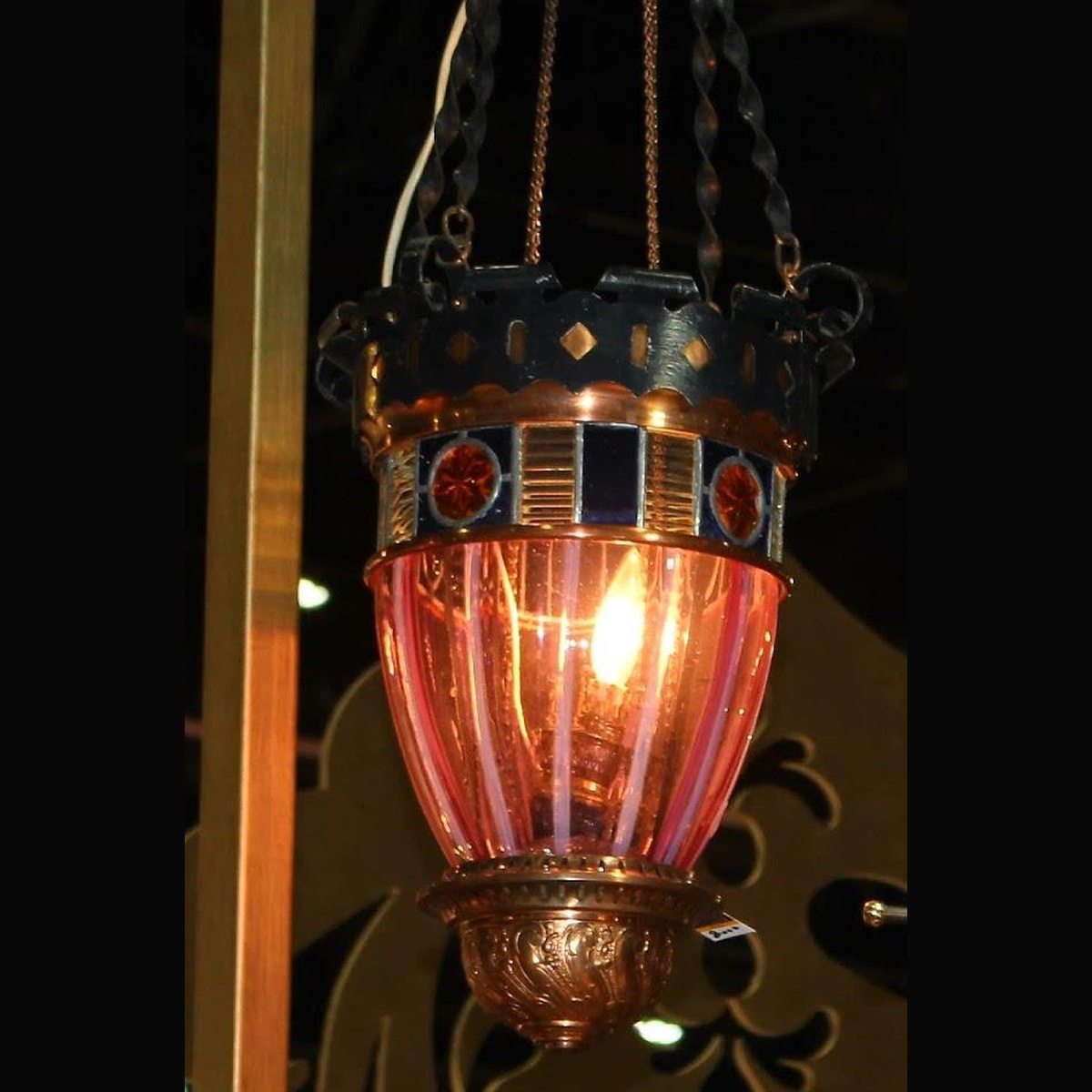 An antique c. 1900 Victorian hanging hall lantern made of ornate copper and brass, featuring a fluted pink cranberry glass body and a border of blue and gold stained glass on a dark background.