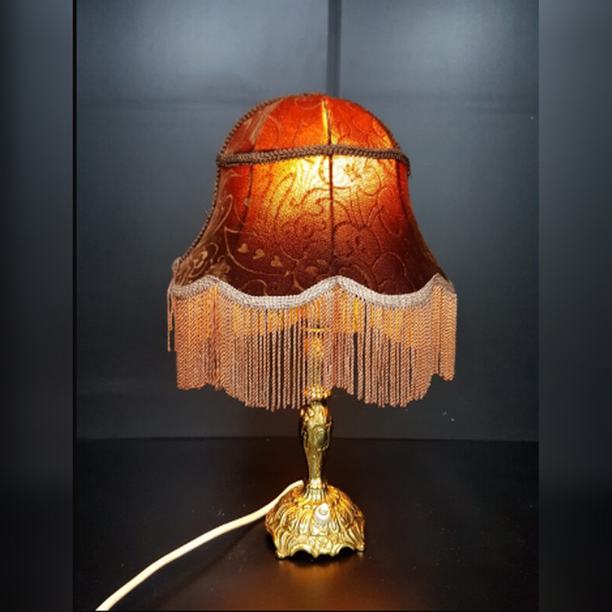 A vintage c. 1950s Victorian-style boudoir table lamp featuring a polished ornate brass base and a glowing red damask brocade fabric shade adorned with dense golden silk fringe.