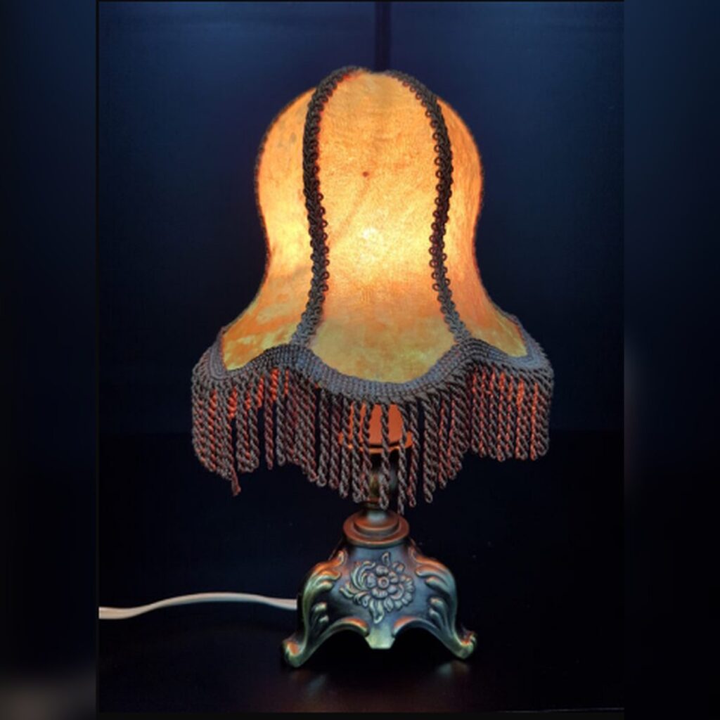 A vintage c. 1950s Art Nouveau style boudoir table lamp with an ornate brass floral relief base and a glowing amber fabric shade decorated with long twisted fringe tassels.