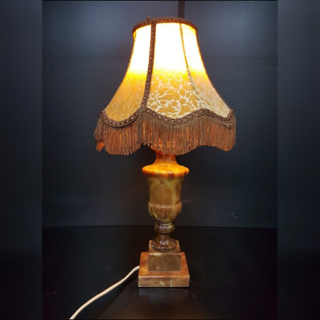 Vintage Neoclassical style table lamp featuring a solid hand-carved brown and amber alabaster stone base with natural veining, topped with a scalloped gold brocade fabric lampshade decorated with long hanging fringe trim.