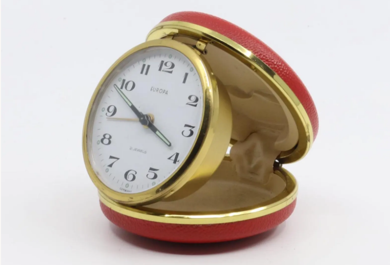 Vintage German mechanical travel alarm clock, c. 1960s, featuring a red folding clam shell case with brass-toned trim, white Arabic numeral dial and luminous-style hands.