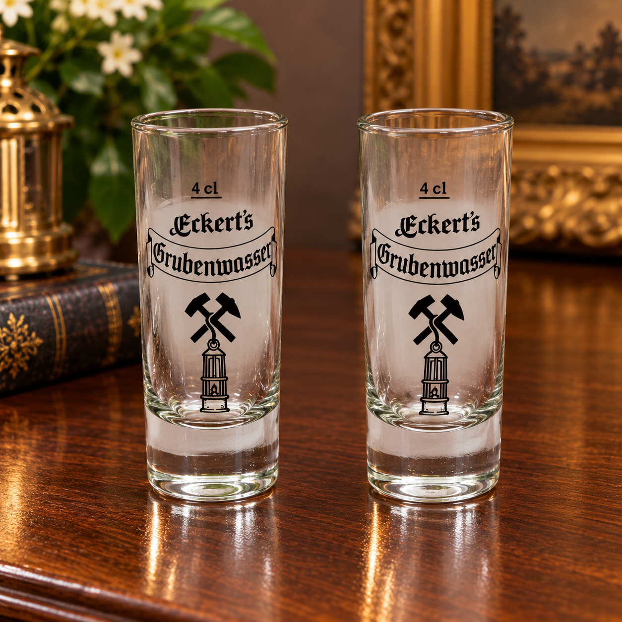 A Pair of vintage German Eckert's Grubenwasser shot glasses with black mining hammer and miner’s lamp emblems, clear glass bodies, and thick bases.